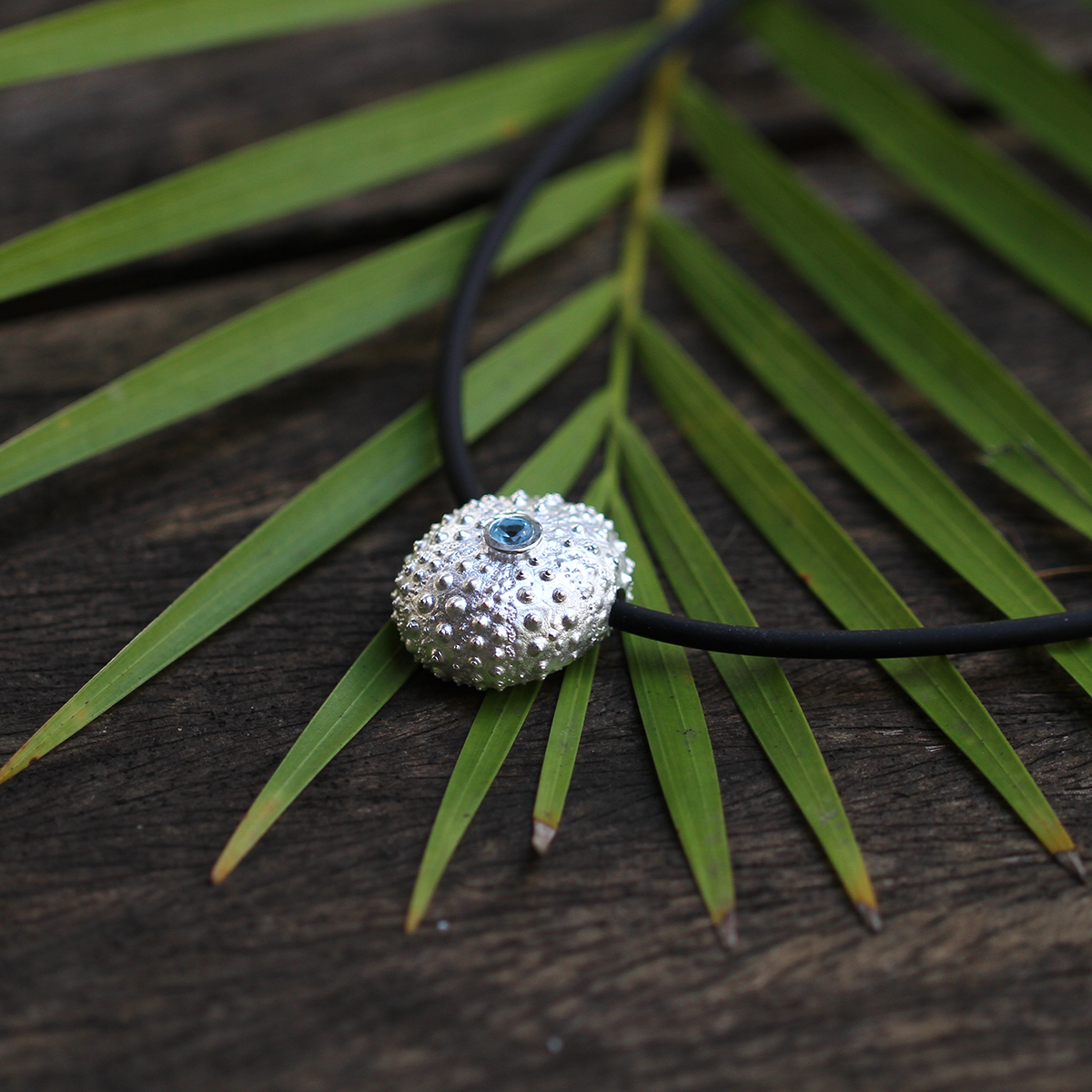 Large, whole sea urchin pendant in sterling silver with a Blue Topas set at its center with a black, rubber chain.