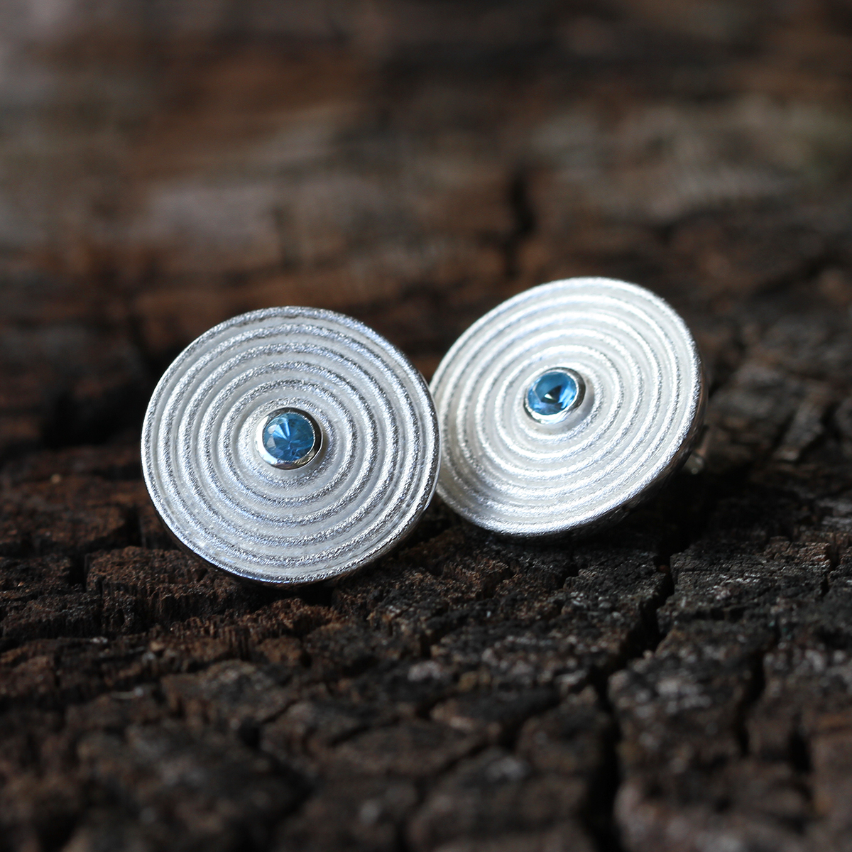 Disc shaped earrings in sterling silver with circular rills originating from Blue stones at the center towards the edge.
